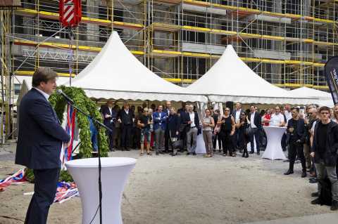 Mann hält Rede bei Feier mit Publikum, Zelten und Baustelle im Hintergrund.