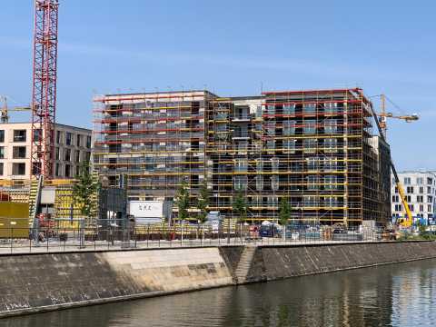 Mehrstöckiges Gebäude mit Gerüst an einem Kanal, Baustellenkräne im Hintergrund.