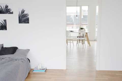 Schlafzimmer mit grauer Bettwäsche, Blick in helles Esszimmer mit Tisch.