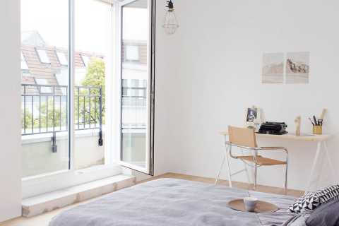 Helles Schlafzimmer mit Schreibtisch, Bett und offener Tür zum Balkon.