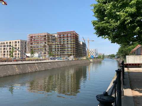 Baustelle mit Gerüst an einem Fluss, Blick auf Kran und Neubauten bei Sonne.