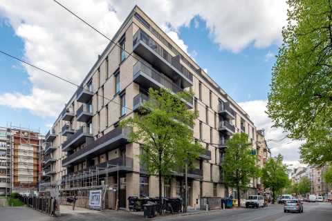Modernes Wohngebäude mit Balkonen an einer Straßenecke, Bäume und Autos davor.