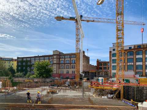 Baustelle mit Kränen, Arbeitern und Gebäuden im Hintergrund bei blauem Himmel.