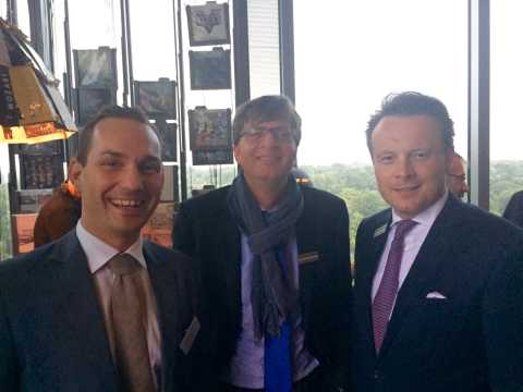Drei Männer in Anzügen lächeln in einem hellen Raum mit Fensterfront.