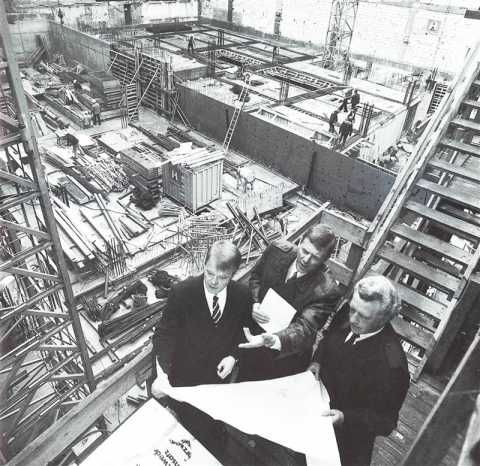 Drei Männer begutachten Baupläne auf einer großen Baustelle mit Arbeitern.