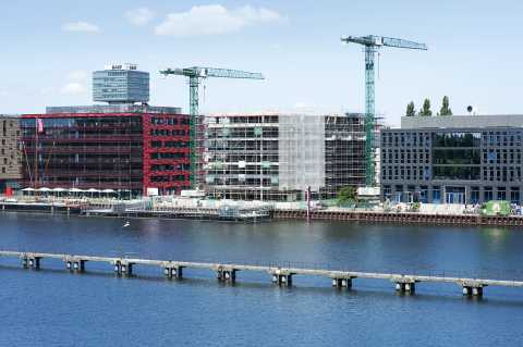 Moderne Gebäude, zwei Baukräne und Fluss mit Steg im Vordergrund.