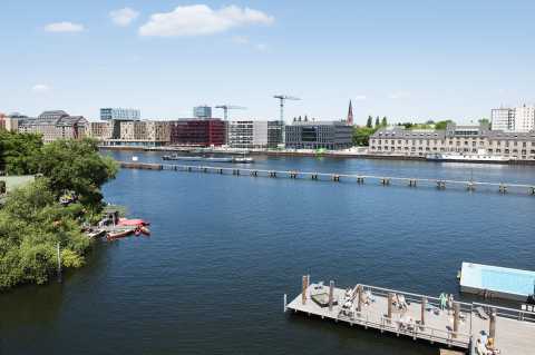 Sonnige Badestelle mit Holzsteg am Fluss, Stadtgebäude im Hintergrund.