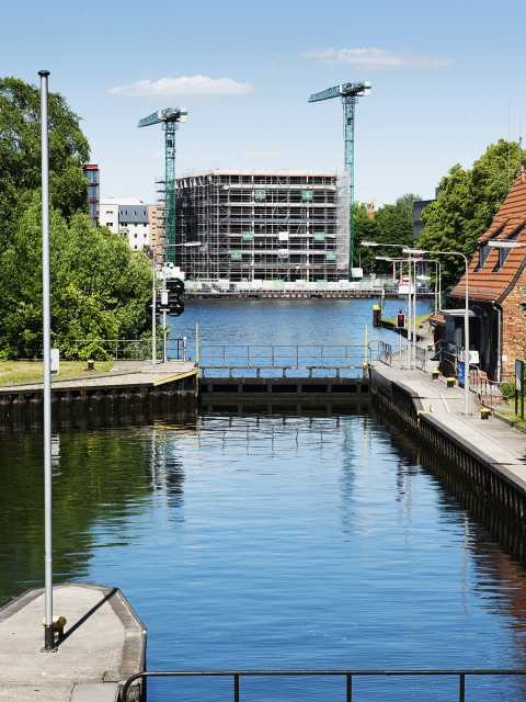 Ein Flusswehr mit ruhigem Wasser vor einer Baustelle und zwei Kränen.