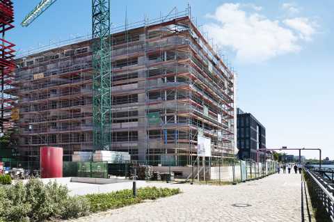 Mehrstöckiges Gebäude im Bau mit Gerüst, Kran und Gehweg am Wasser.