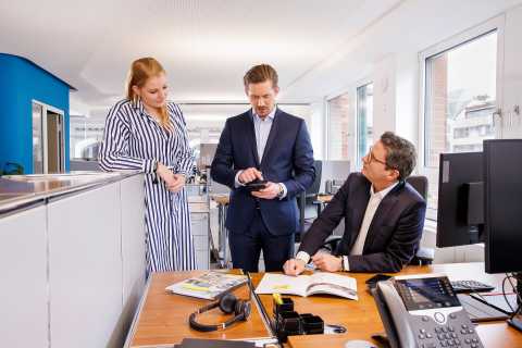 Drei Personen im Büro sprechen zusammen, einer sitzt am Schreibtisch.