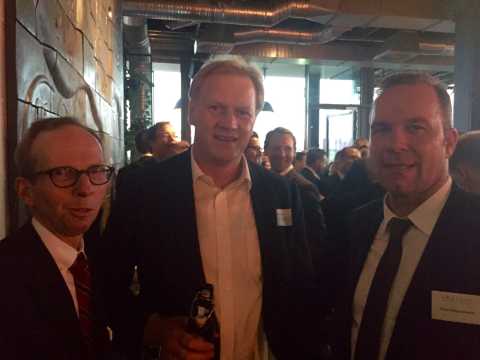 Drei Männer in Anzügen bei einer Veranstaltung in einem modernen Raum.