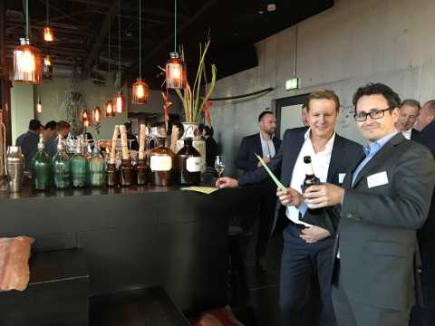 Zwei Männer im Anzug stehen an einer Bar mit Flaschen und unterhalten sich.