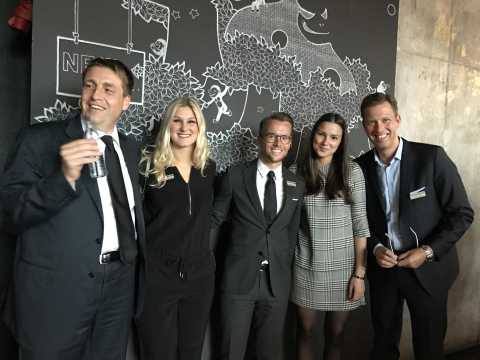 Fünf Menschen in Businesskleidung lächeln vor einer Wand mit Tierzeichnung.