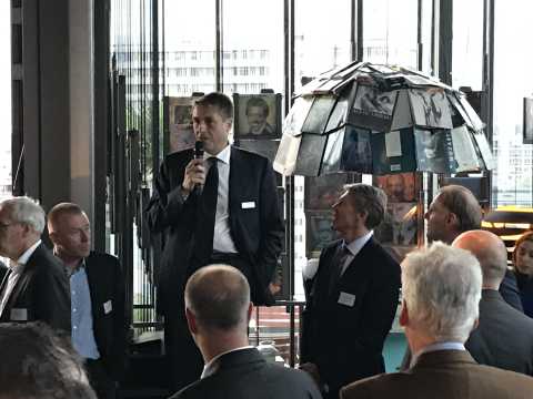 Mann im Anzug spricht mit Mikrofon vor einer Gruppe bei einer Veranstaltung.