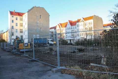 Baustelle mit Bagger und Zaun, dahinter mehrstöckige Wohnhäuser.