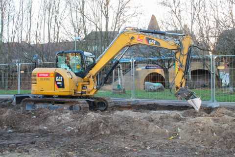 Gelber Bagger auf Baustelle hebt Erde aus, im Hintergrund ein Zaun und Bäume.