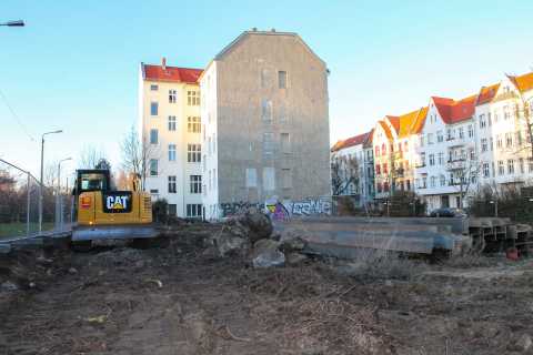 Bagger auf Baustelle vor Wohnhäusern, mit Baumaterial und grauem Gebäude.