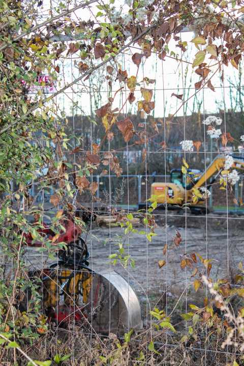 Baumaschine hinter Maschendrahtzaun mit Pflanzen im Vordergrund.