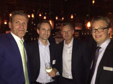 Vier Männer in Anzügen stehen zusammen in einer Bar und lächeln.