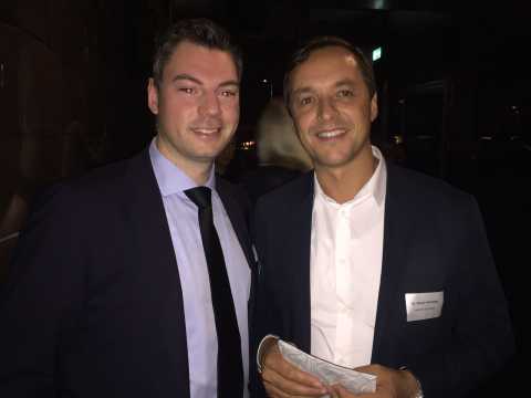 Zwei Männer im Anzug lächeln in dunkler Umgebung in die Kamera.