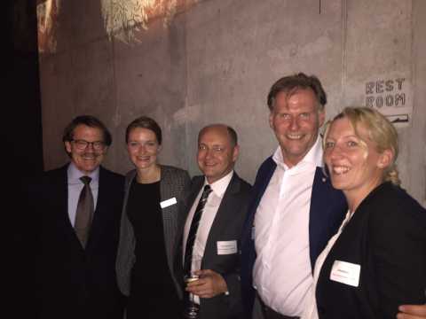 Fünf Menschen in Businesskleidung posieren lächelnd vor einer Betonwand.