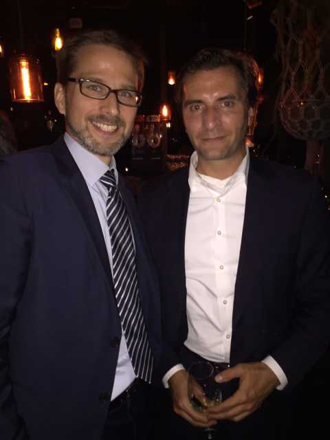 Zwei Männer im Anzug stehen in einer Bar, einer hält ein Weinglas.