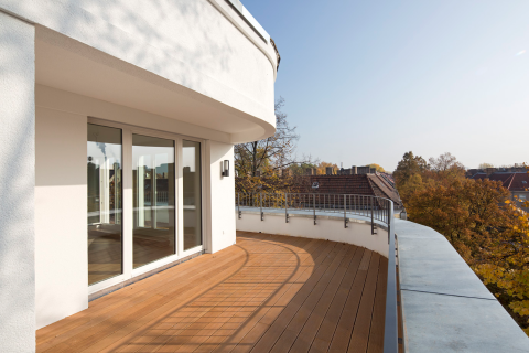 Großer Balkon mit Holzterrasse, Glasfront und Blick auf Dächer und Bäume.