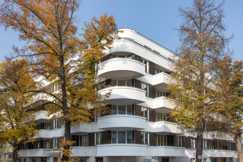 Weiße Wohnhausfassade mit runden Balkonen, umgeben von herbstlichen Bäumen.