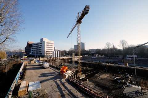 Große Baustelle mit Kran, Baugrube, Baumaterial und Gebäuden im Hintergrund.