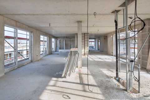 Innenraum einer Baustelle mit großen Fenstern, Rohbau und offenen Leitungen.
