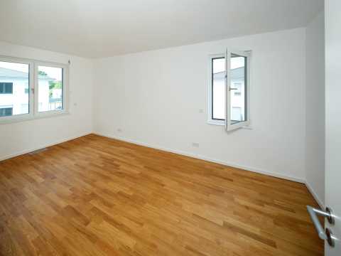 Leeres Zimmer mit Parkettboden, weißen Wänden und zwei geöffneten Fenstern.