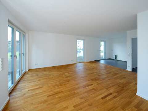 Leerer, heller Raum mit Parkettboden und großen Fenstern, weiße Wände.