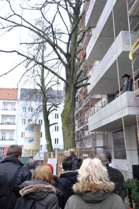 Menschen stehen vor einer Baustelle mit Baum und Gerüst, ein Mann auf dem Balkon.