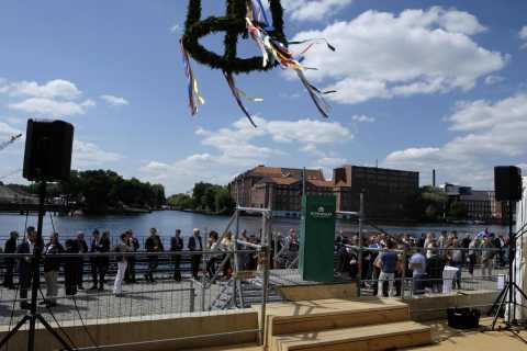 Menschen feiern ein Richtfest am Wasser, Kranz mit Bändern hängt über Bühne.