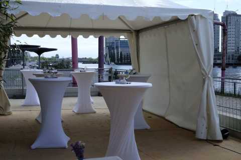 Stehtische mit weißen Hussen unter Pavillon an einem Flussufer, dahinter Skyline.