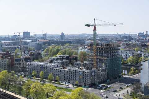 Baustelle mit Kran und Rohbau in einer Stadt, umgeben von Bäumen und Straßen.