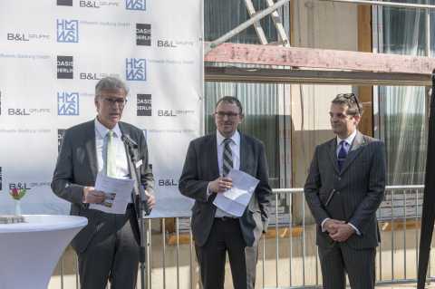 Drei Männer im Anzug stehen bei einer Rede vor einer Baustelle und Werbewand.