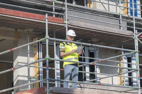 Bauarbeiter mit Helm und Weste steht auf Gerüst, hält Tablet in der Hand.