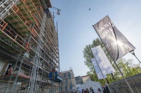 Baustelle mit Gerüst, Kran, Fahnen und Menschen bei sonnigem Wetter.