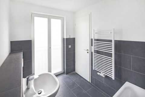 Modernes Bad mit dunklen Fliesen, Waschbecken, Tür, Fenster und Handtuchheizkörper.