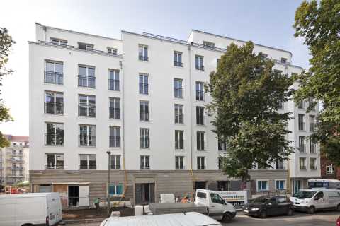 Mehrstöckiges, modernes Wohnhaus mit weißen Fassaden, davor Bäume und Autos.