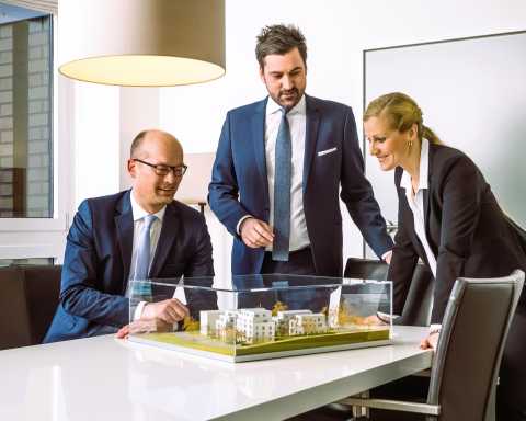 Drei Personen im Anzug betrachten ein Architekturmodell auf einem Tisch.