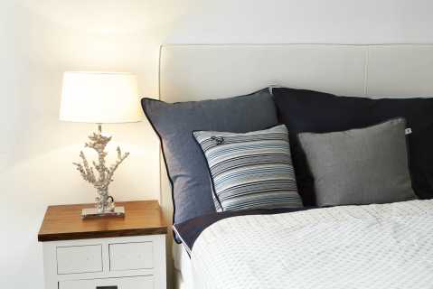 Nachttisch mit Lampe, daneben ein Bett mit mehreren Kissen und heller Bettdecke.