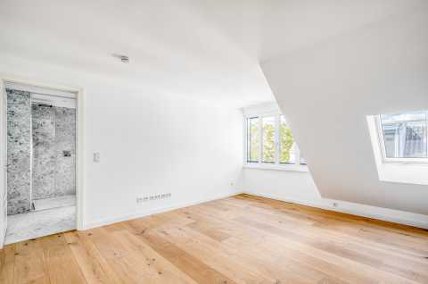 Helles, leeres Zimmer mit Holzfußboden, Fenstern und Zugang zum Bad.