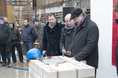 Mehrere Männer in Winterkleidung bei einer Grundsteinlegung auf einer Baustelle.