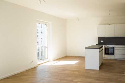 Helle, leere Wohnung mit offener Küche, Holzboden und Balkonzugang.