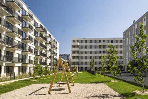 Moderner Wohnhof mit Spielplatz, Schaukeln und jungen Bäumen an sonnigem Tag.