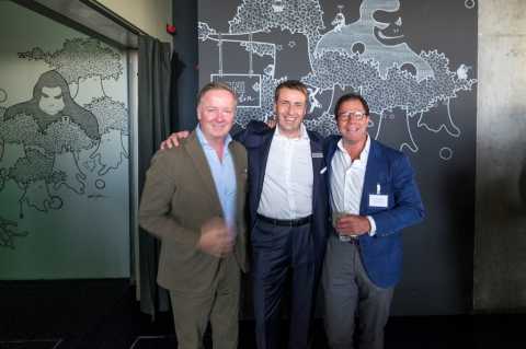 Drei Männer in Anzügen posieren zusammen vor einer Wand mit Illustrationen.