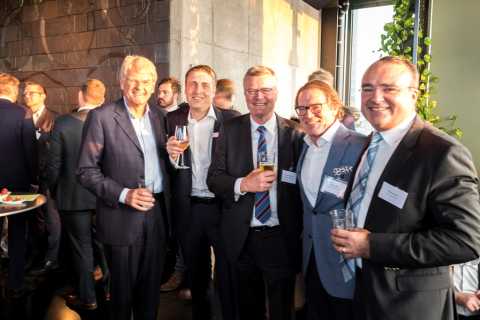 Fünf Männer im Anzug lächeln bei einer Veranstaltung mit Getränken in der Hand.