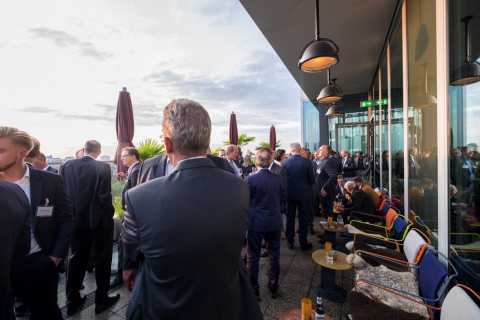Viele Menschen in Anzügen unterhalten sich auf einer Terrasse bei Sonnenuntergang.